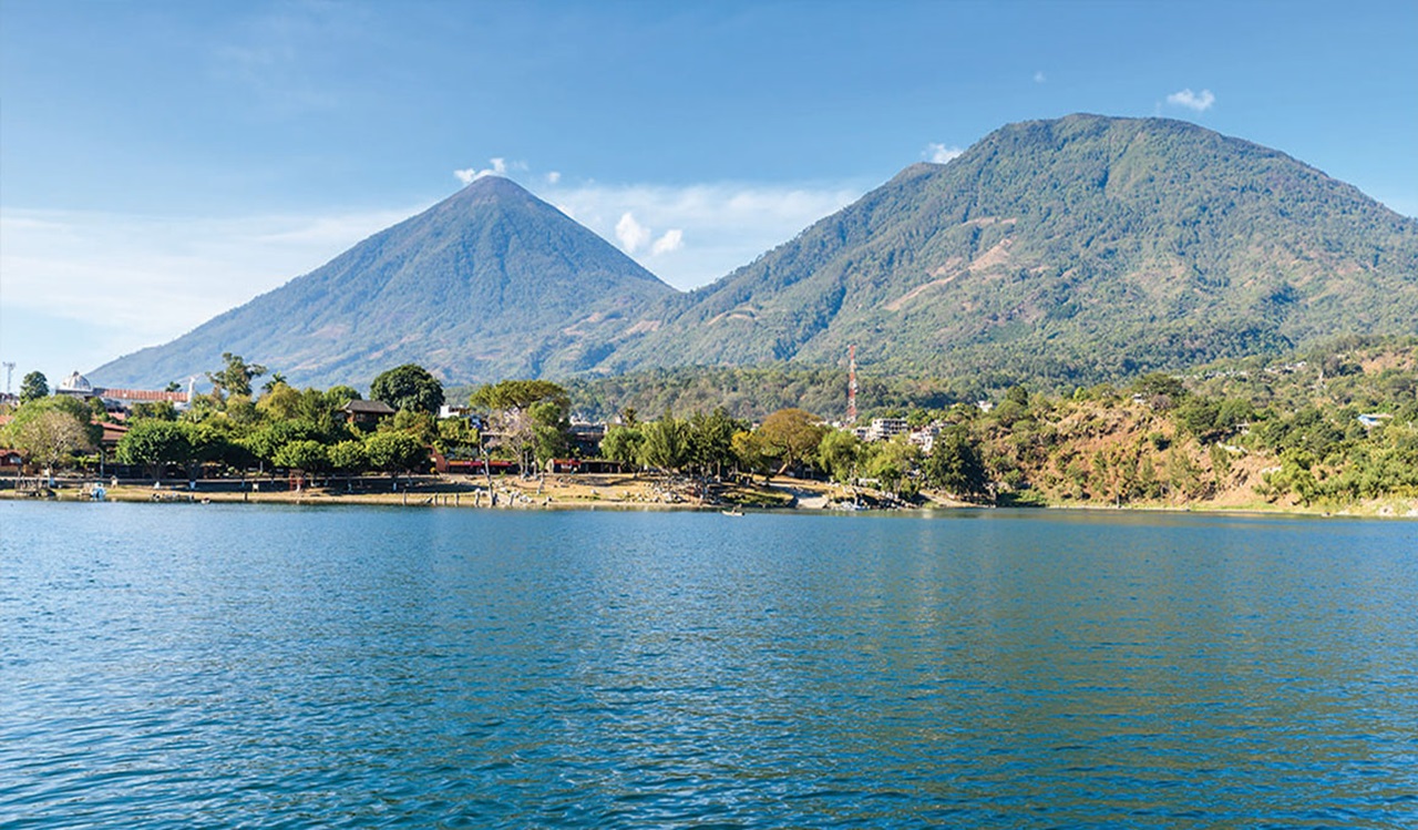 Guatemalan landscape with ocean water in the forefront, and mountains, trees and buildings in the background.