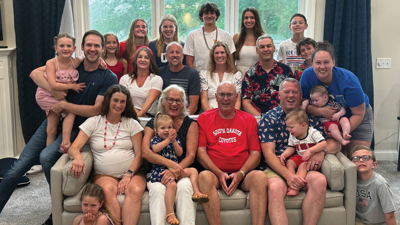 The Schuneman's pose for a picture with their children and grandchildren, sitting on a couch in front of a big window with natural lighting.