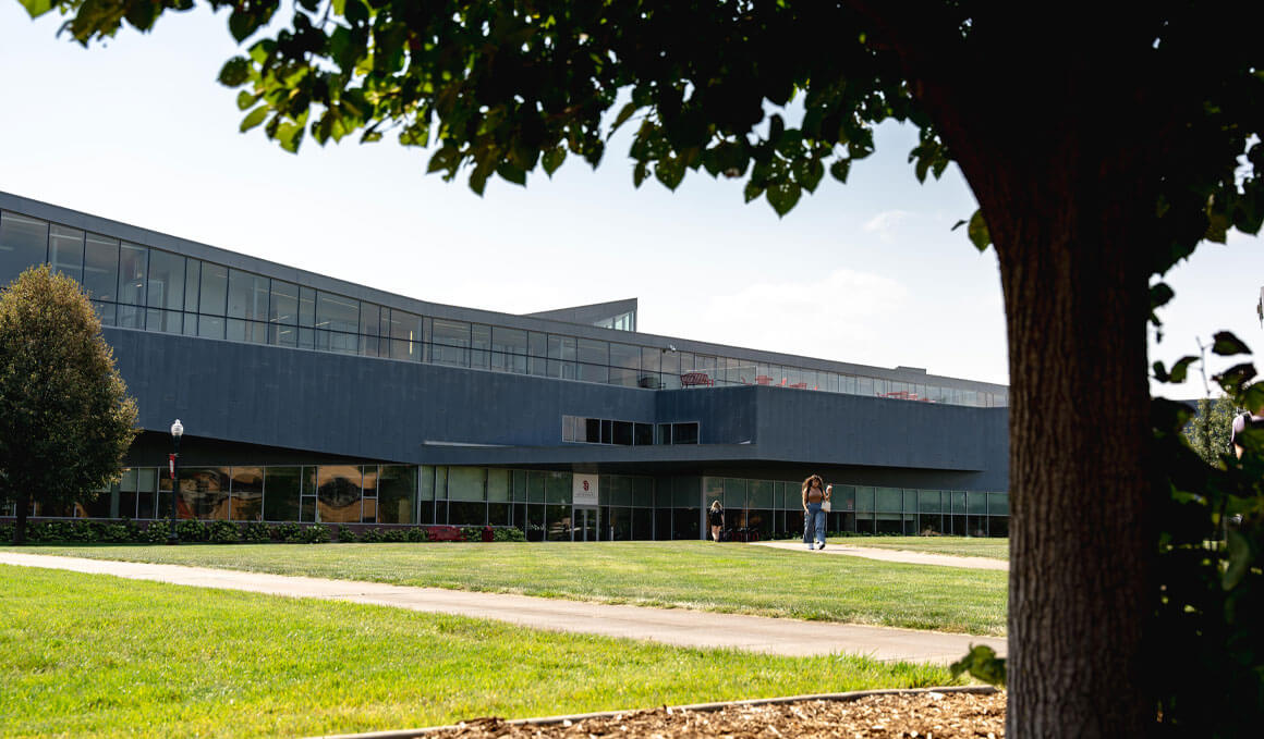 The Beacom School of Business sits on a sunny USD campus. There are a few students in the foreground, along with trees and green grass.