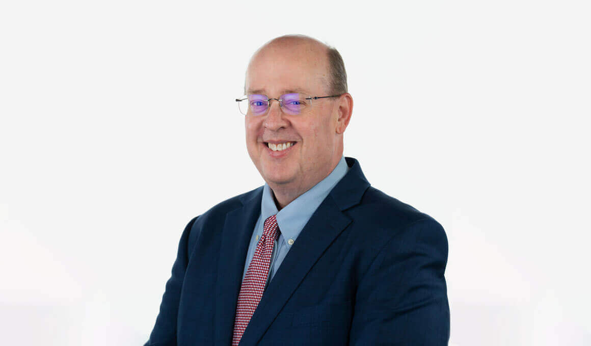 Matt Fairholm sits his professional photo, which is against a white background. He is wearing a suit and tie, glasses and smiling at the camera.