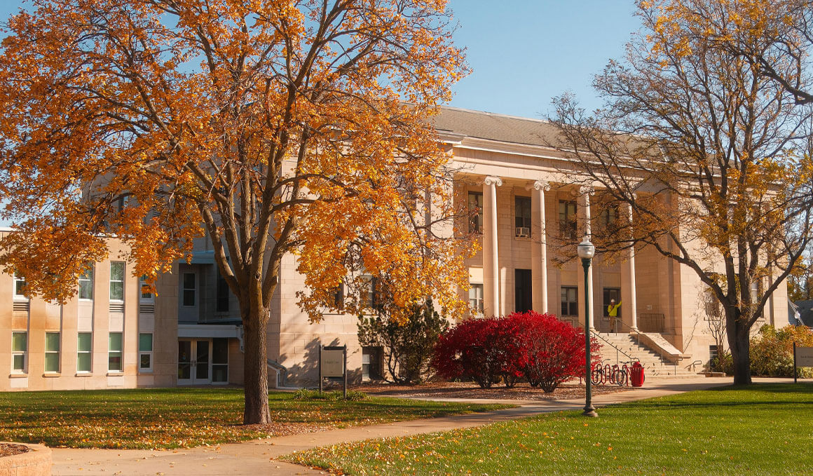 The USD College of Arts & Sciences stands behind orange autumn trees and red shrubs, creating a calm, welcoming scene on a sunny day.