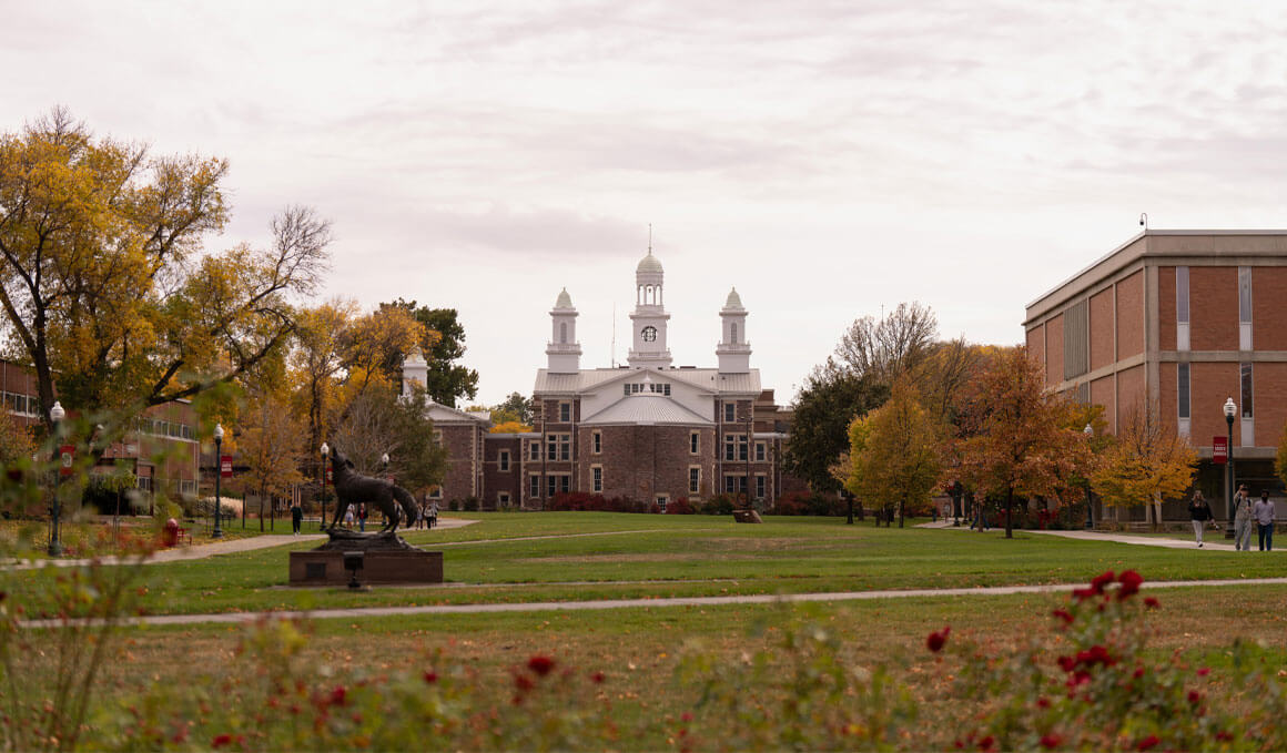 A front view of the USD campus, with Old Main and other historic university buildings in the background, along with the Coyote Legacy statue. In the forefront, fall leaves, bushes and flowers can be seen.
