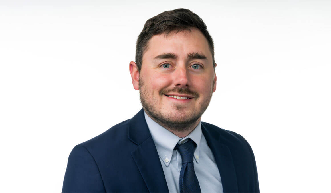 Thomas Mrozla poses for his professional photo against a white backdrop. He is wearing a blue suit and tie and smiling at the camera.