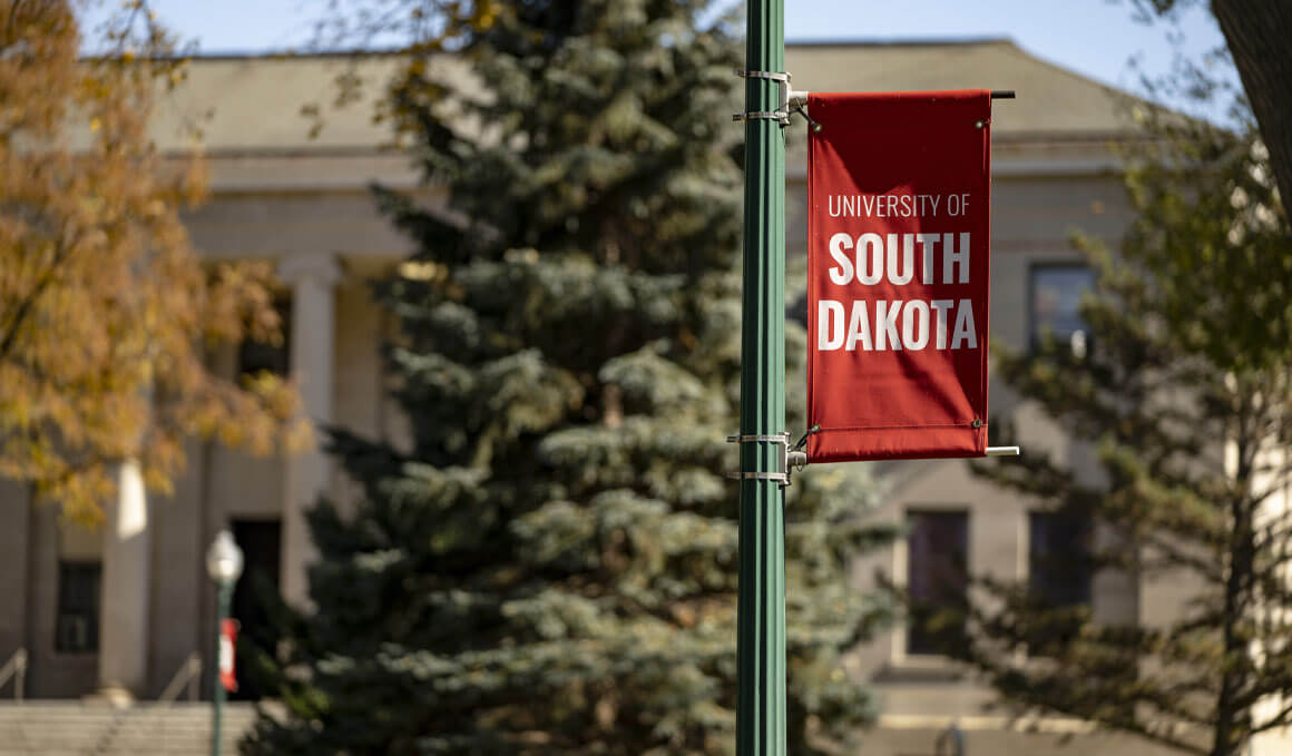 A red University of South Dakota banner hangs from a green campus lamppost, with an academic building and evergreen trees softly blurred behind it.