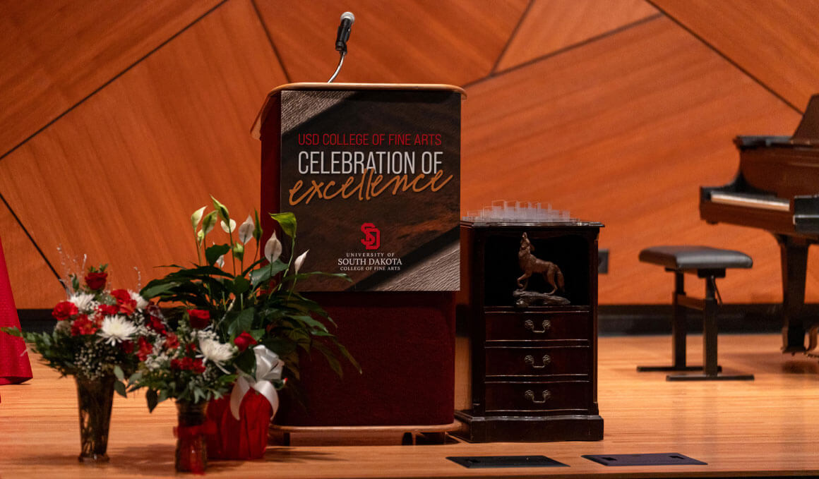 A view of the stage in the Colton Recital Hall, which is adorned with flowers, a podium and awards, at the Warren M. Lee Center for Fine Arts at the University of South Dakota. A grand piano is also visible in the background.