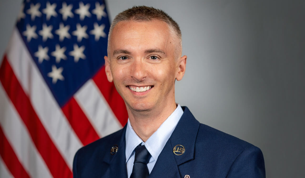Brian Mann stands in his U.S. service member uniform and poses in front of an American flag and gray backdrop.