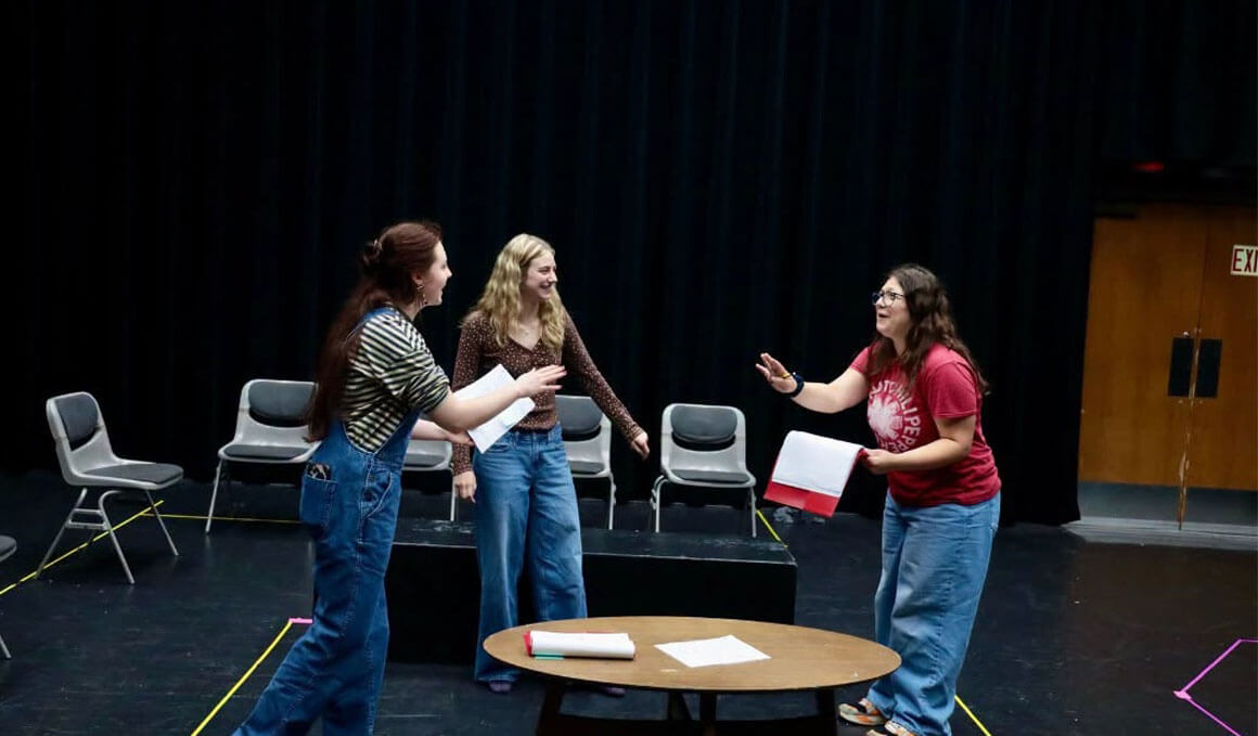 Three USD students rehearse on a stage, gesturing around a table with papers and a book; chairs and a black curtain frame the casual, collaborative scene.