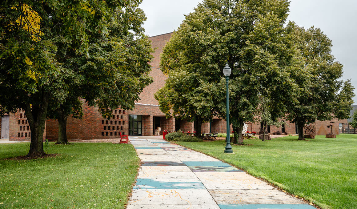 The entrance of the College of Fine Arts' building is shown, surrounded by an overcast sky and trees, along with a walking path that leads to the doors and is covered in fallen leaves.