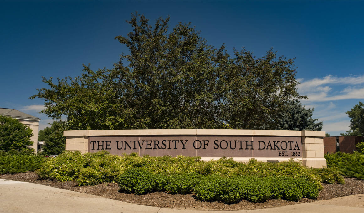 The University of South Dakota sign sits on the USD campus. It is made out of brick and surrounded by green bushes and trees, along with a blue, cloudy sky.