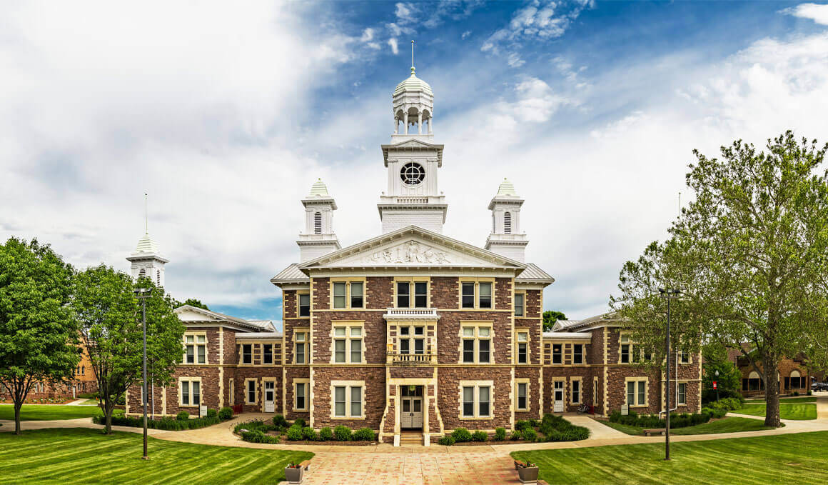 USD's historical Old Main sits on the university's campus. It is surrounded by blue, cloudy skies, green trees and grass.