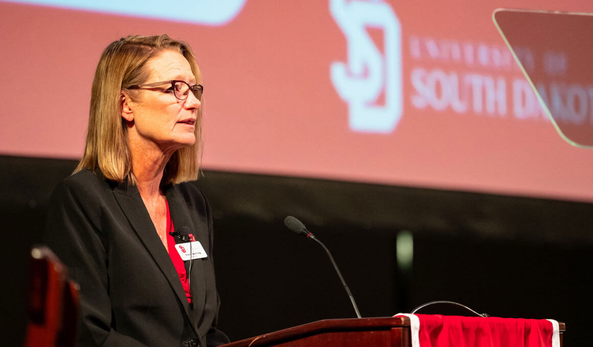 USD President Sheila K. Gestring stands at a podium, delivering the 2026 State of the University address, with a PowerPoint on the screen behind her.