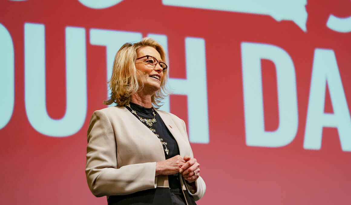 USD President Sheila K.Gestring stands on stage in front of a red screen with oversized white lettering, smiling and speaking to an audience.
