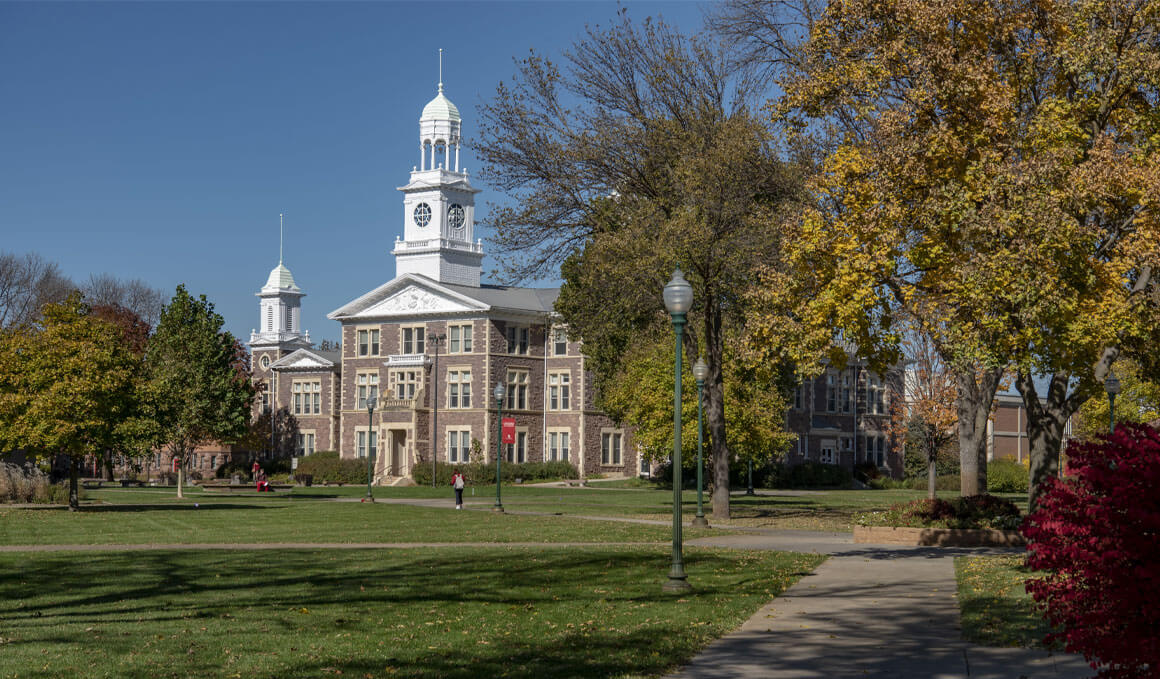Old Main sits on the USD campus as students walk on the sidewalk in front of it. In the forefront are bushes that are starting to turn to their fall colors, along with leaves lightly falling on the grassy area.