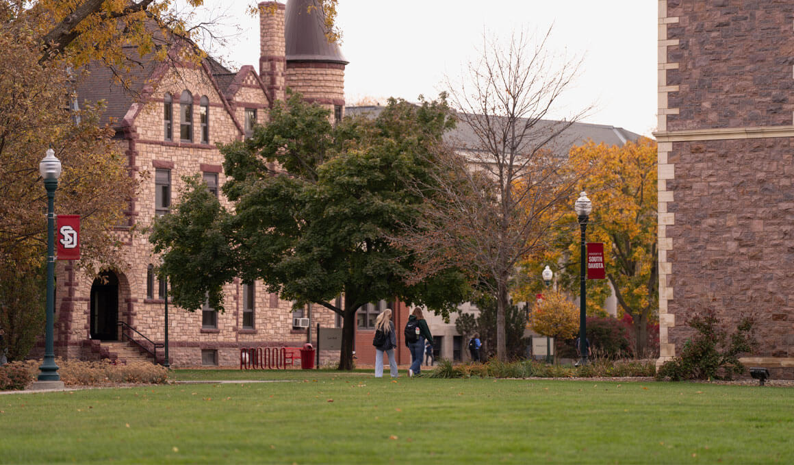 USD students walk along a paved pathway on campus and a  historic stone building and autumn foliage can also be seen.