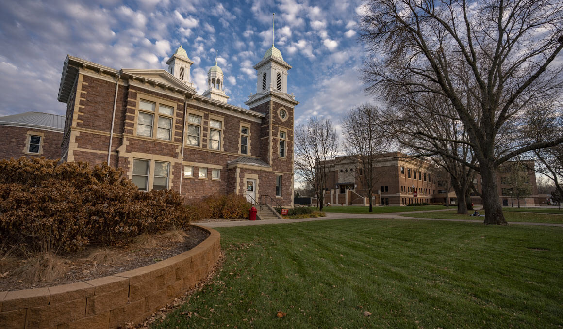 Old Main sits beside a broad green lawn and bare trees under a dramatic, cloud-streaked sky.