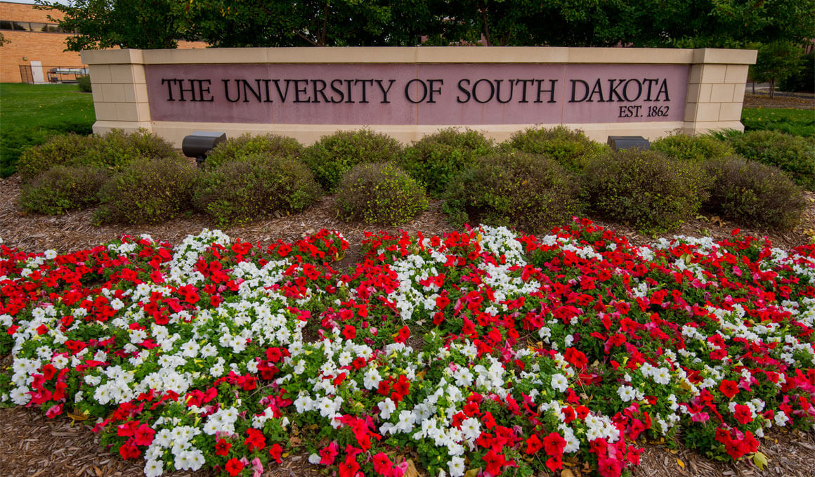 A flower bed full of red and white flowers sits in front of a stone and brick "University of South Dakota" sign on the USD campus.