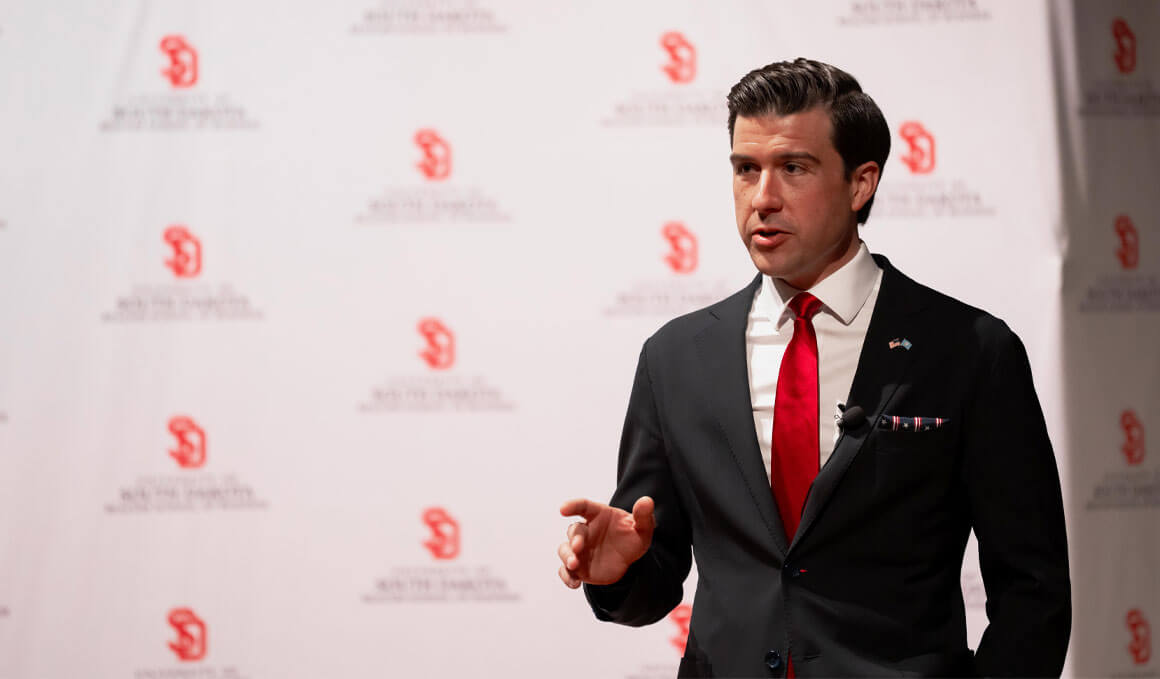 Luke Lindberg stands on stage in front of a USD branded backdrop. He is wearing a black suit and bright red tie.