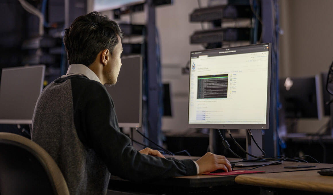 A student is working at a computer/desktop station. His back is turned to the camera and the computer monitor is displaying a white screen with a black coding box on it.