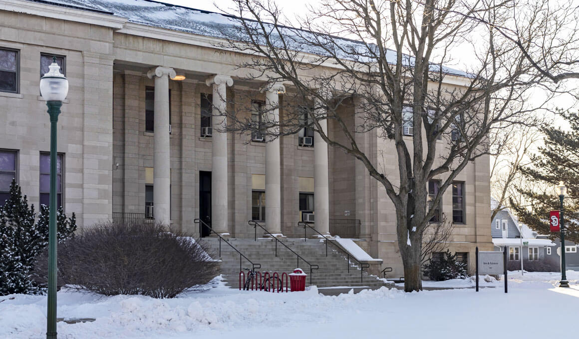 The College of Arts & Sciences sits on a wintry USD campus, with snow and lamp posts in the foreground.