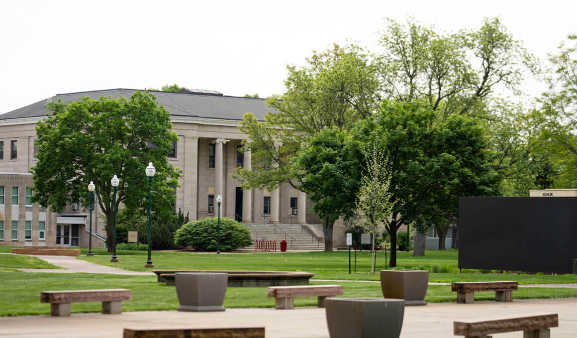 A faraway view of the College of Arts & Sciences on the USD campus. It is surrounded by green lawn and trees, with light poles, benches and sidewalk in the foreground.
