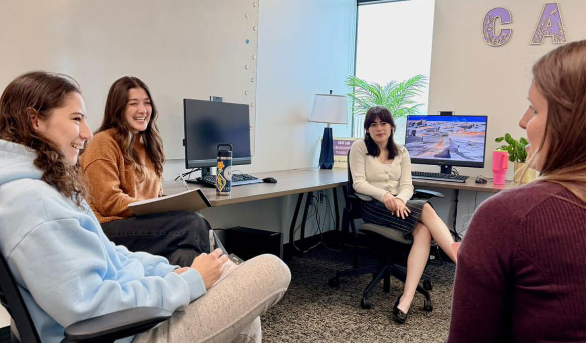 Dr. Danzi and her team of student researchers sit in the CAST Lab having a discussion. They are surrounded by computer monitors, along with a window and plants.