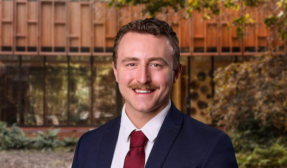 Caleb Swanson, a 2024 USD Alumn, poses for his professional photo. In the background, there is a building with windows and some trees and plants on the right hand side.