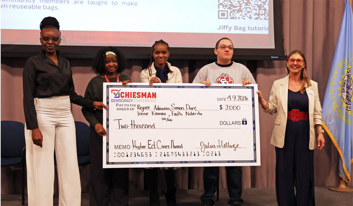 Winners of the inaugural Higher Education Civic Engagement Award stand on stage with Julia Hellwege. They are holding a check and smiling at the camera. 