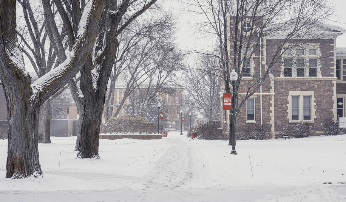 An old building sits on a snowy USD campus, with trees in the foreground and lightpoles that are holding red USD banners.