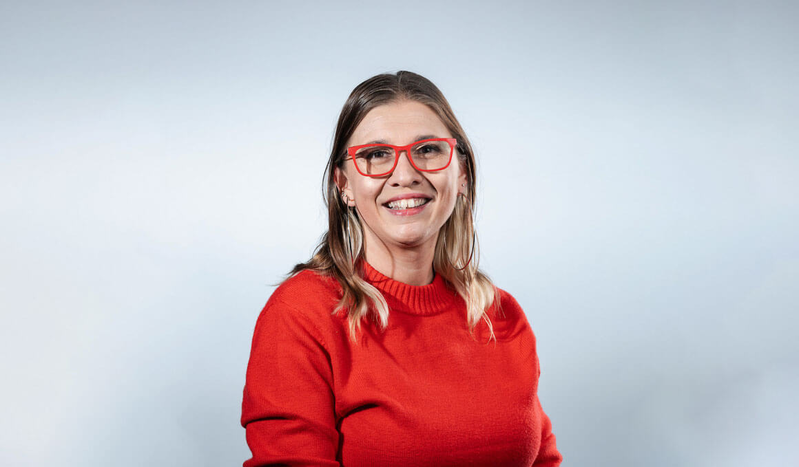 Julia Hellwege, Ph.D., is sitting on a stool, posing for her professional photo. She is wearing a bright red sweater and bright red glasses.