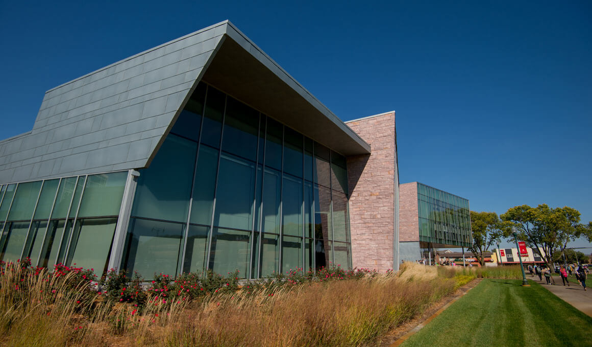 The Muenster University Center sits on the University of South Dakota campus, surrounded by plants and green grass on a sunny day. Students are walking on the sidewalk in front of the building.