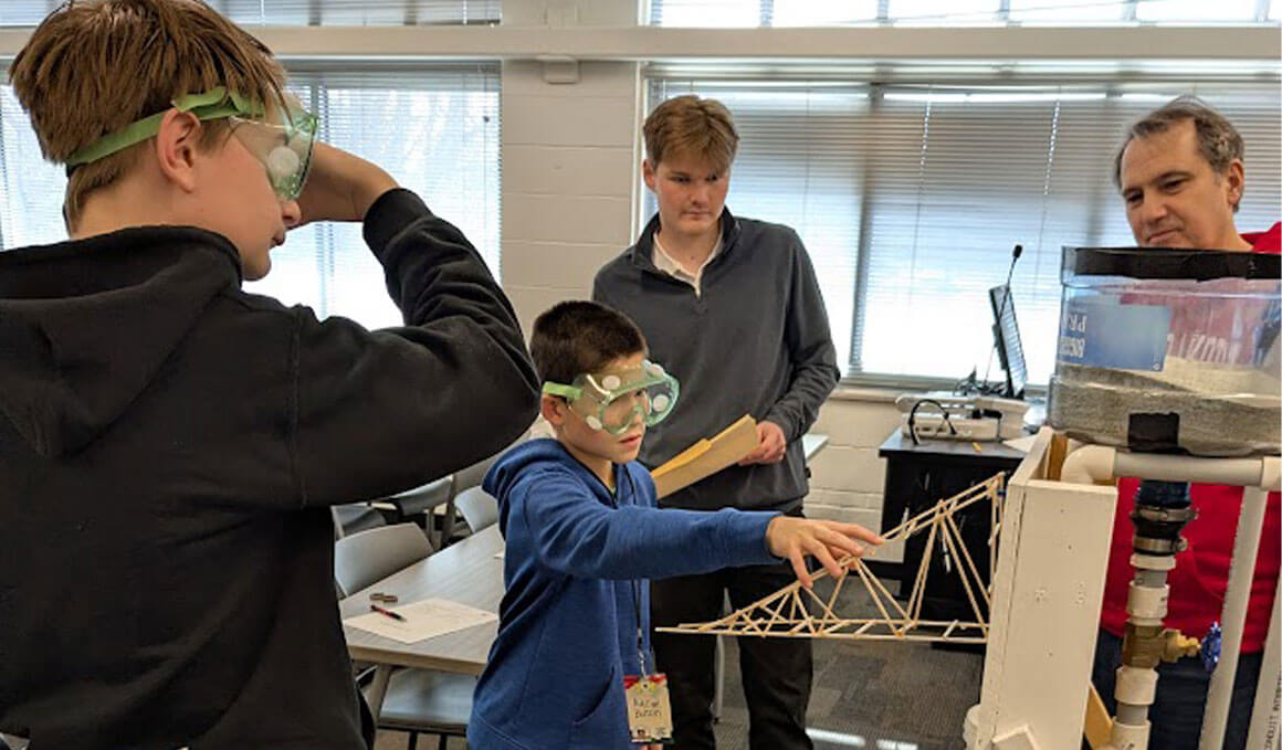 A child wearing safety goggles reaches toward a small wooden bridge model being tested under a load in a classroom at the South Dakota Science Olympiad competition.