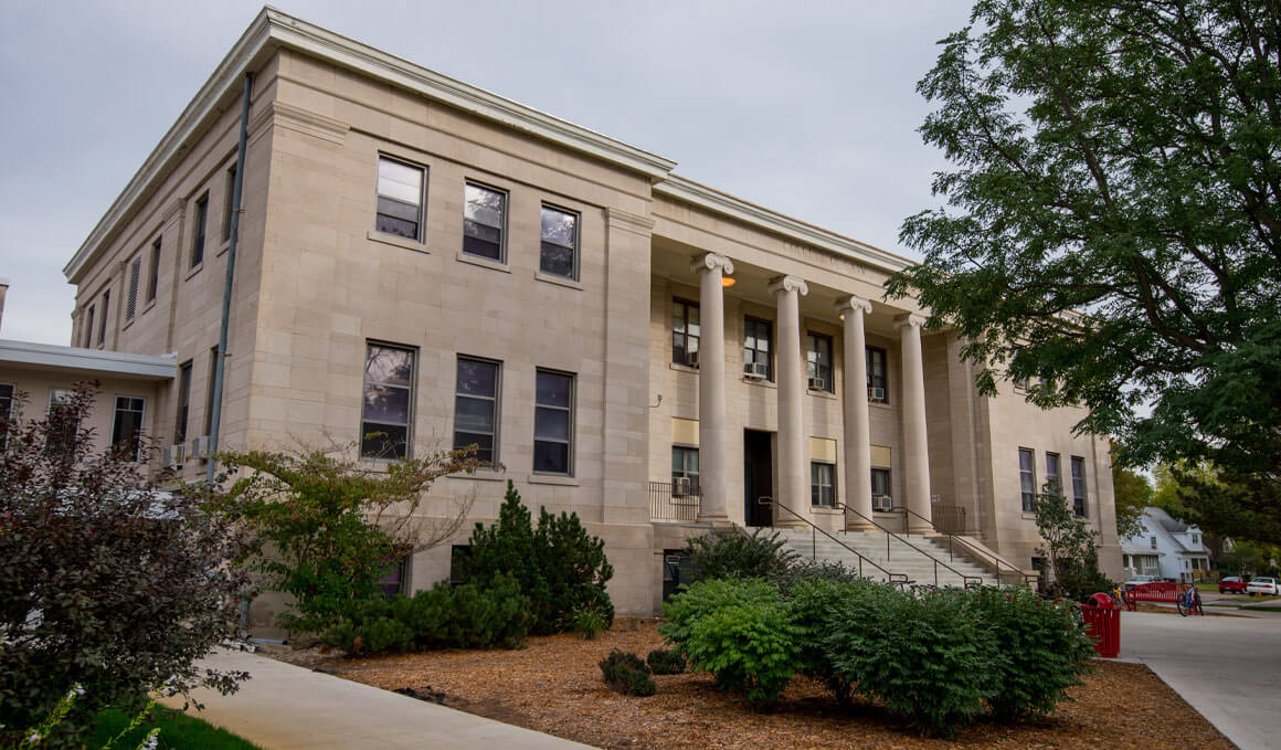 The College of Arts & Sciences exterior, showing a two-story stone building with tall columns at the front entrance, with steps leading up to the doors and landscaped trees, along with shrubs, surrounding the building.