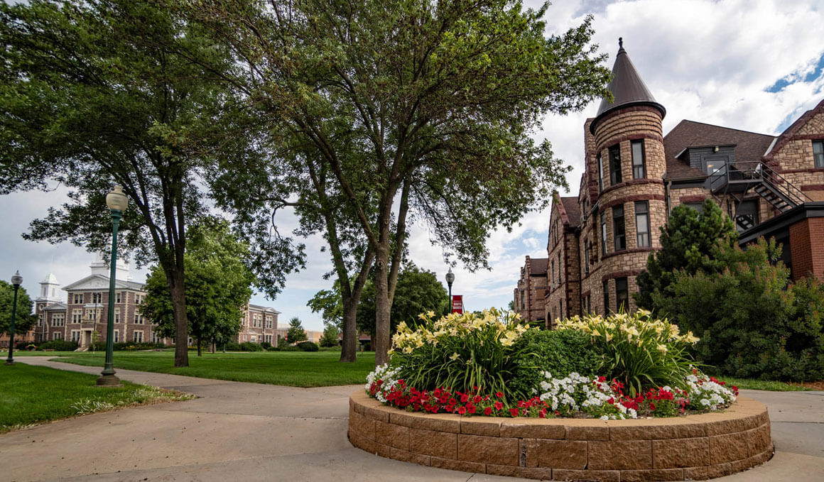 A tree-lined campus walkway surrounds a circular flower bed in front of historic stone buildings, including one with a turret, under a partly cloudy sky.