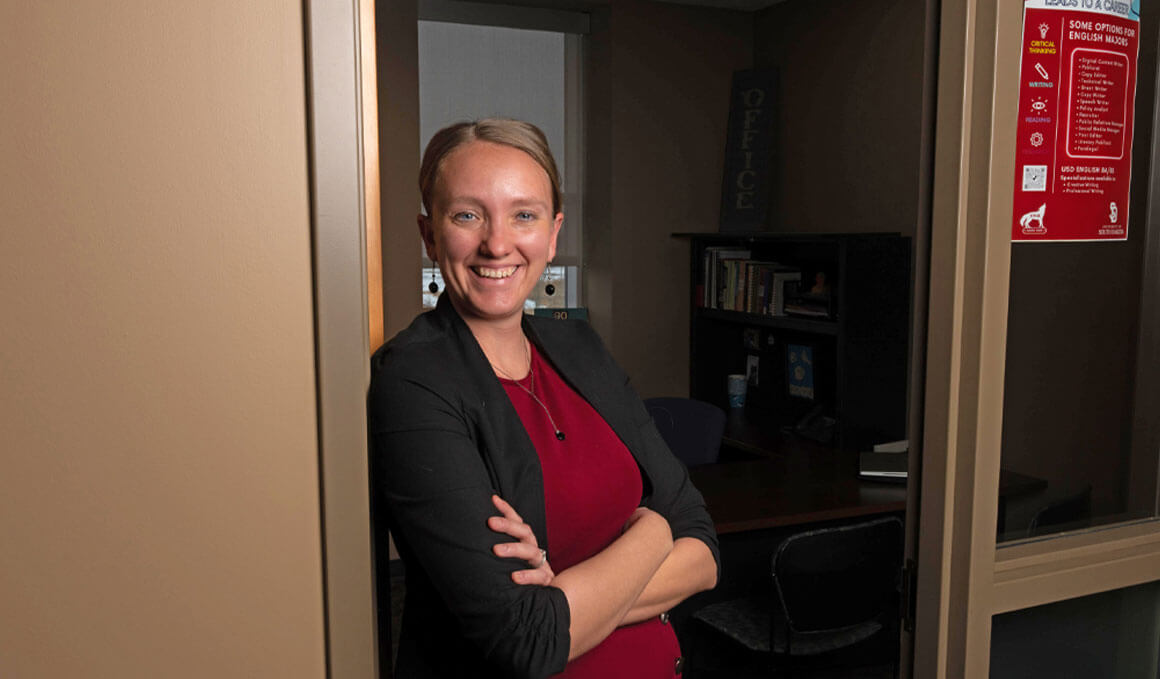  Cheyenne Marco, Ph.D., a senior lecturer in the USD Department of English, stands in front of her office door. She is wearing a red dress with a black cardigan and smiling at the camera.