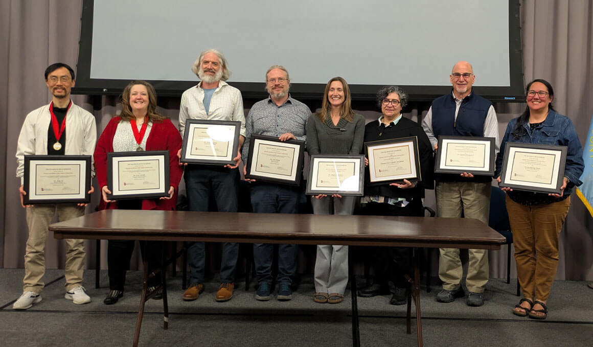 A group of USD College of Arts & Sciences faculty stand on a stage, in front of a curtain and screen, holding their framed awards. There is a table in front of them and they are smiling while looking at the camera.