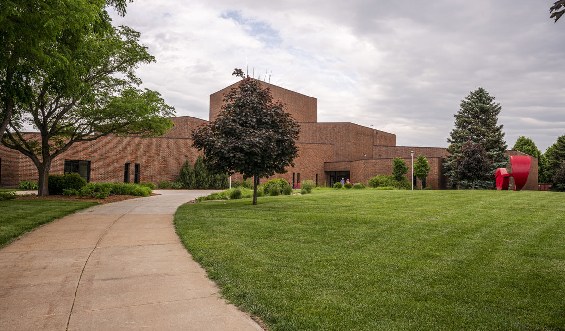 The exterior of the College of Fine Arts sits on an overcast USD campus, with a green lawn in the foreground, and a red statue to the right of the building.