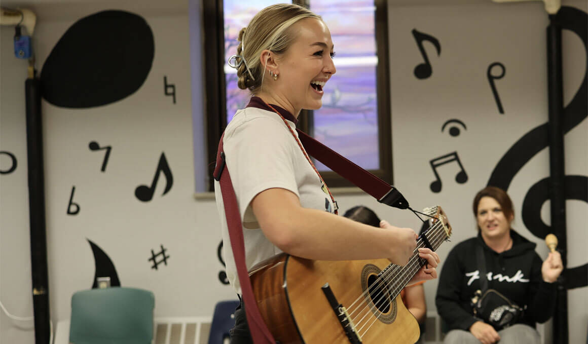 Kari Edman, a USD alumna, stands in a music room at her job. She is smiling and playing a guitar. A participant can be seen sitting in the background. The walls of the room are filled with cutouts and pictures of music related symbols and items.