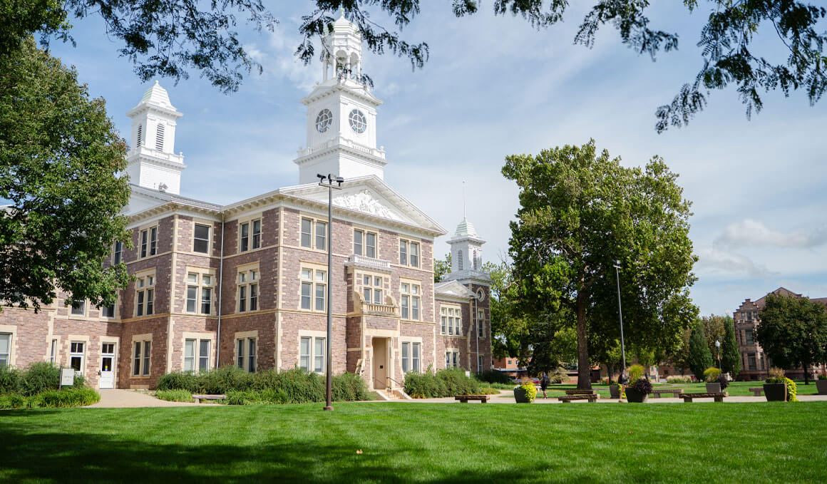 Old Main sits on a sunny USD campus, surrounded by green grass and trees.
