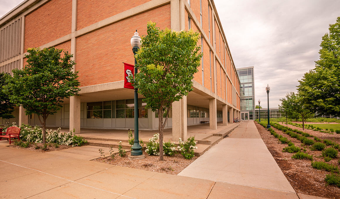 Outside of I.D. Weeks library on USD Vermillions campus. Sidewalks and trees with light posts and USD banners.