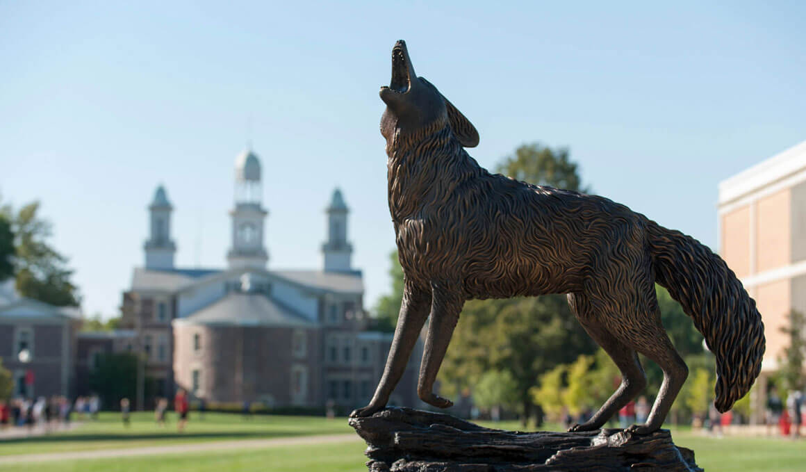 The Coyote Legacy statue sits in the foreground on the USD campus, with Old Main and students visible in the background.