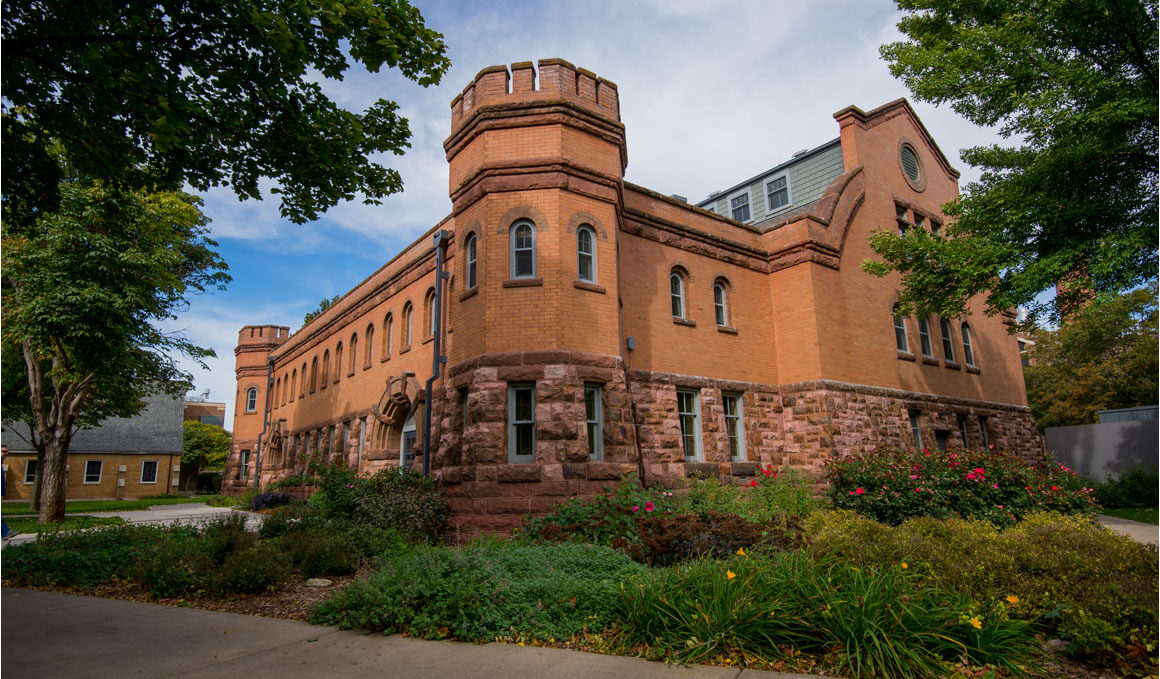 The Belbas Center sits on the USD campus, with green plants and colorful flowers in the forefront. It is surrounded by green trees and a blue sky with white clouds.