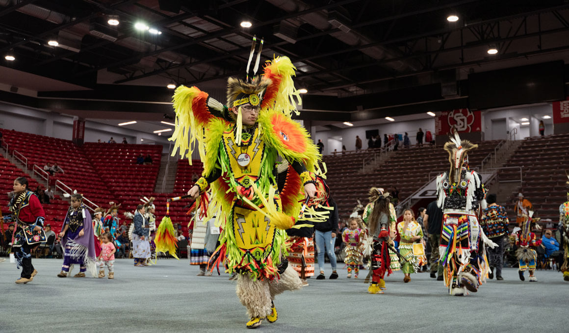 This image shows a Wacipi dancer in bright neon yellow and orange traditional regalia, performing in the DakotaDome. The dancer is surrounded by other participants in colorful attire with stadium seating in the background.