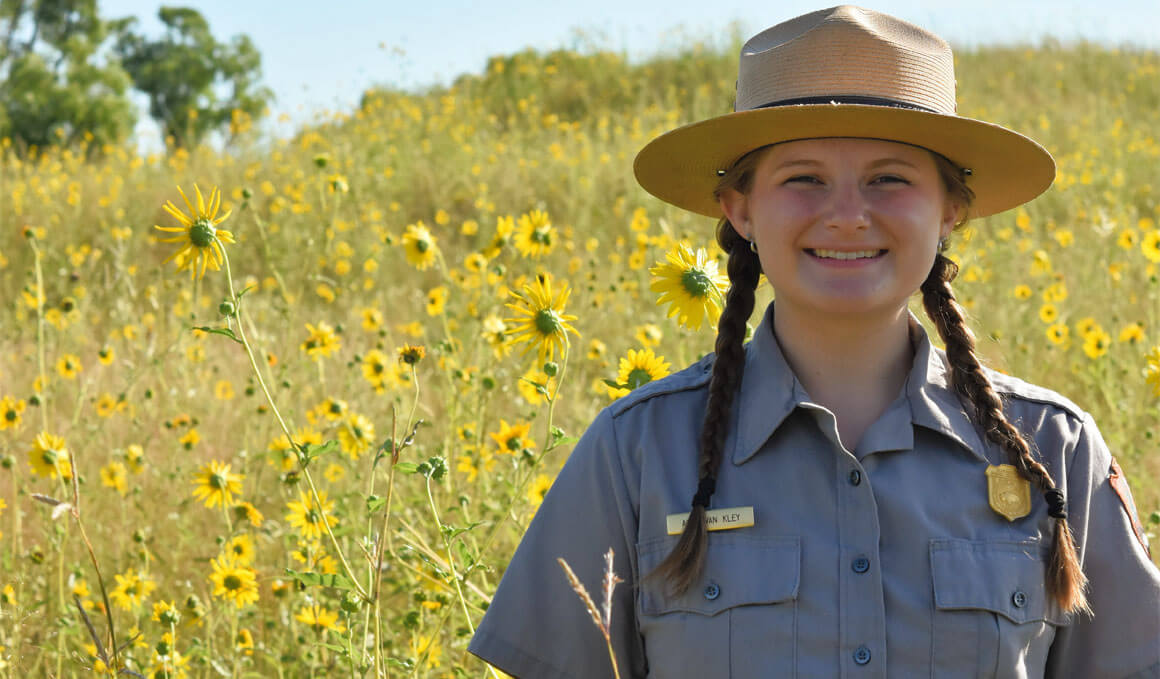 Anna Van Kley smiles at the camera while standing in a field of wildflowers. She is wearing her park ranger gear and hat.