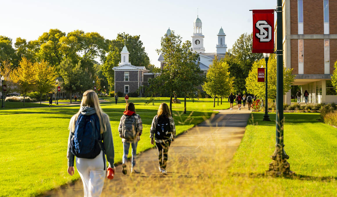 Students walk with backpacks towards Old Main. They are surrounded by green grass and other USD campus buildings, along with light poles that have USD banners attached.