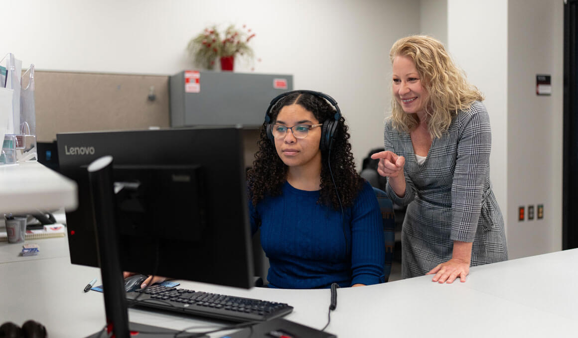 Angela Jackson stands in the Office of Digital Accessibility with a colleague, who is seated, pointing toward a computer monitor with a smile. The seated woman has curly dark hair, wears glasses and a headset, and is focused on the screen.