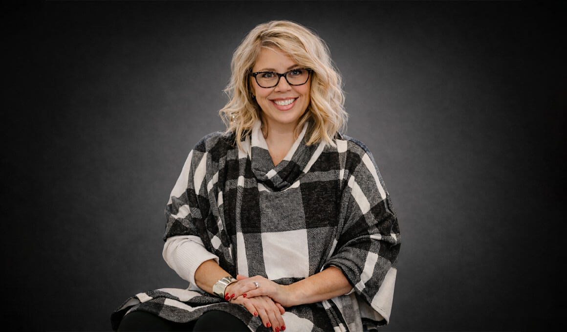 Emily Quinn sits on a stool and poses for her professional headshot against a black background. She is wearing a black and white checkered shirt, glasses and smiling at the camera.