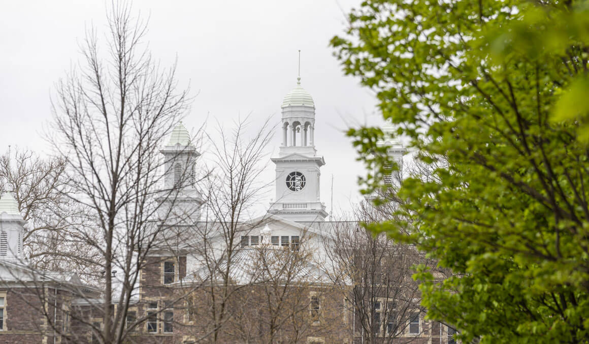 Old Main rises in the background, partially framed by leafy green branches and bare trees on a gray, overcast day.