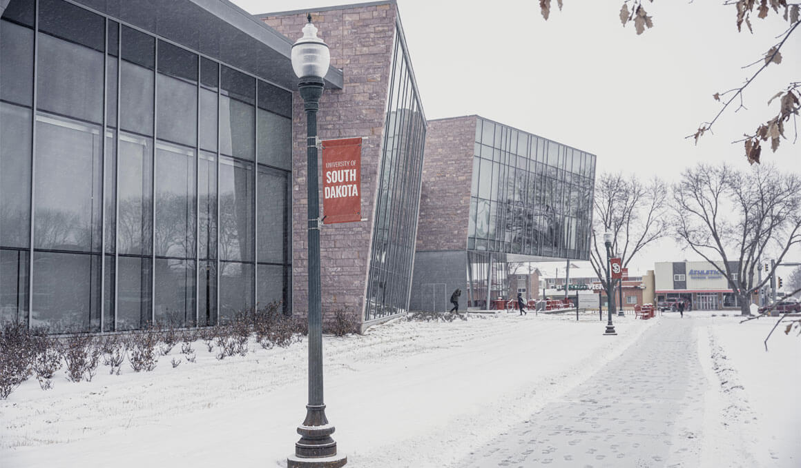 A photo of the Muenster University Center with snow surrounding it. A light pole is in the forefront with a red USD banner attached to it. 