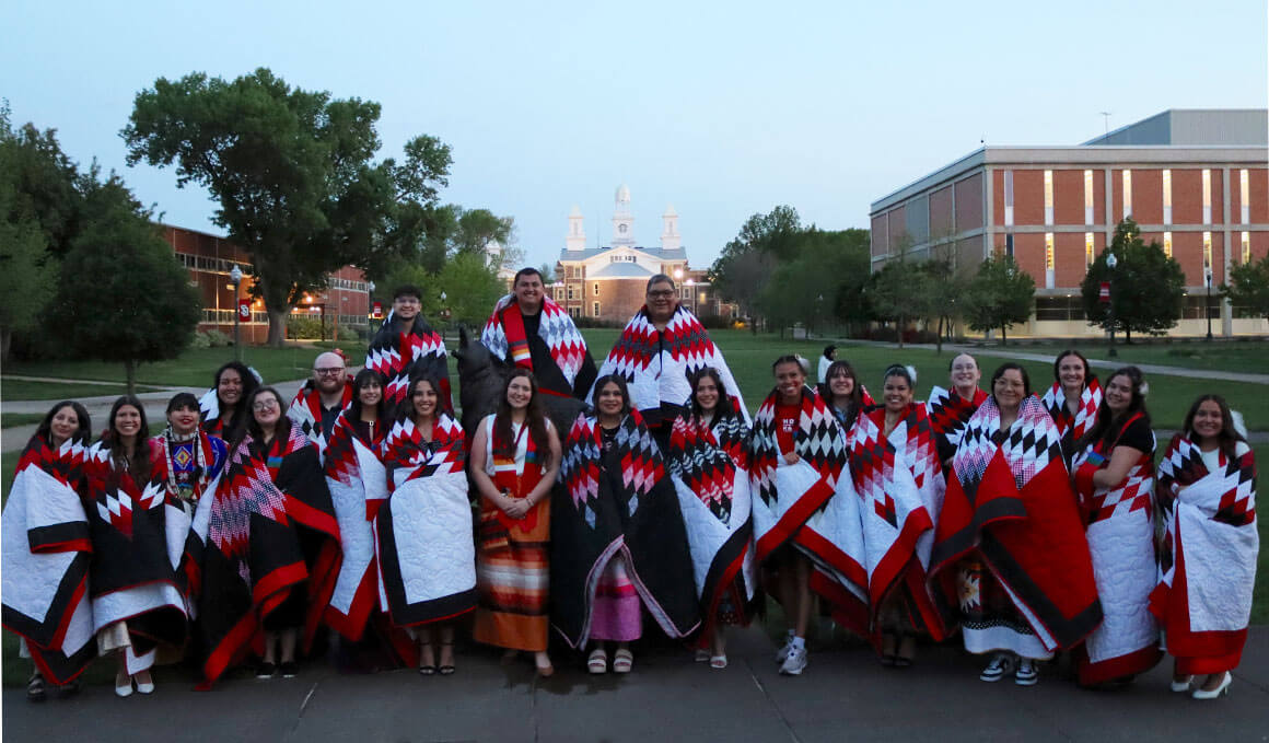 A group of people wearing red, black and white patterned blankets stand together outdoors on the USD campus at dusk with buildings and trees in the background.