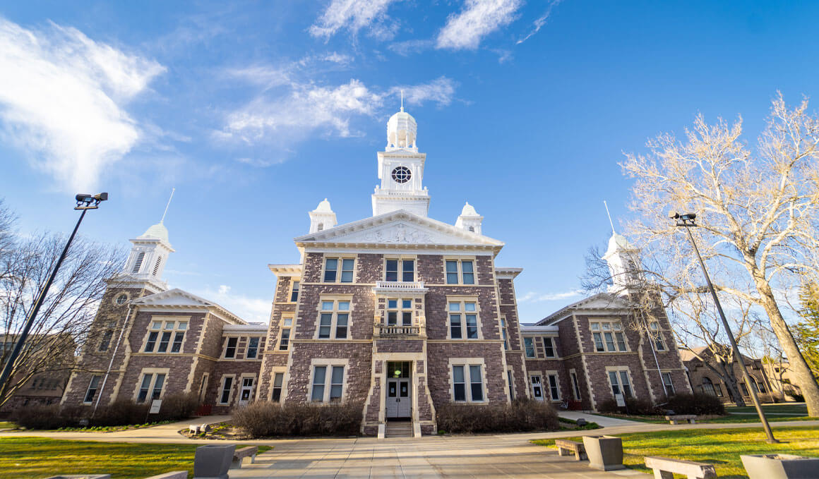 A close up photo on the exterior of Old Main, sitting on the USD campus, with a sunny blue sky and white clouds in the background, along with green grass and trees in the forefront.